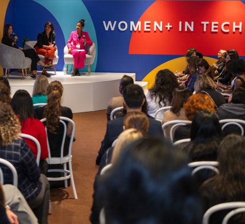 A seated crowd listens as three speakers have a panel conversation on stage at a Women+ in Tech event