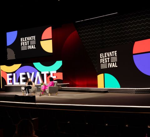 Two people sitting on chairs on a stage and talking at a past Elevate Festival