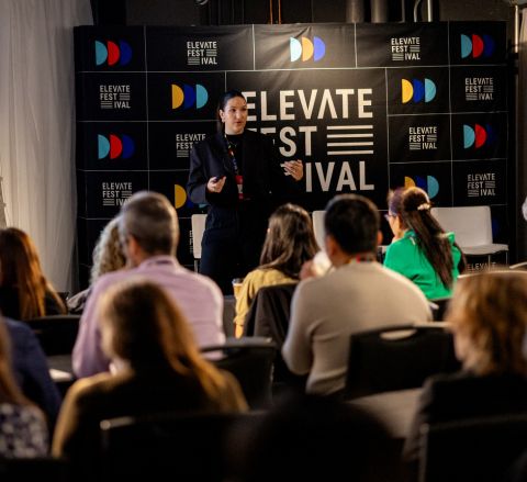 A person standing on a stage speaking to a seated audience at a past Elevate Festival